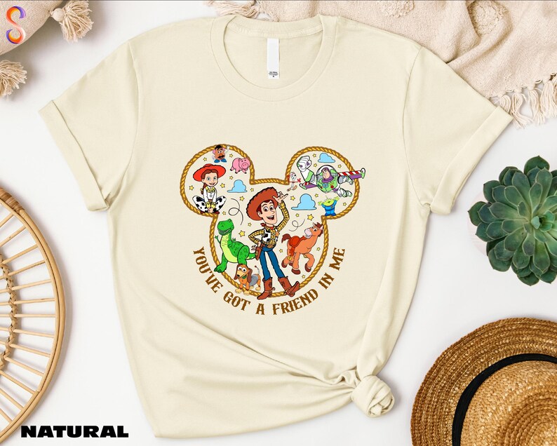 A beige t-shirt with a cartoon character design and text, along with various other items such as a hat, a woven basket, and a succulent plant.