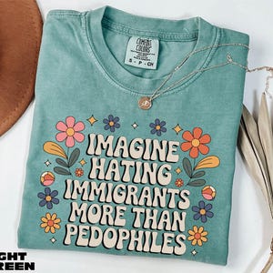 A turquoise t-shirt with a colorful graphic design and text, along with a necklace and some dried leaves, on a white background.