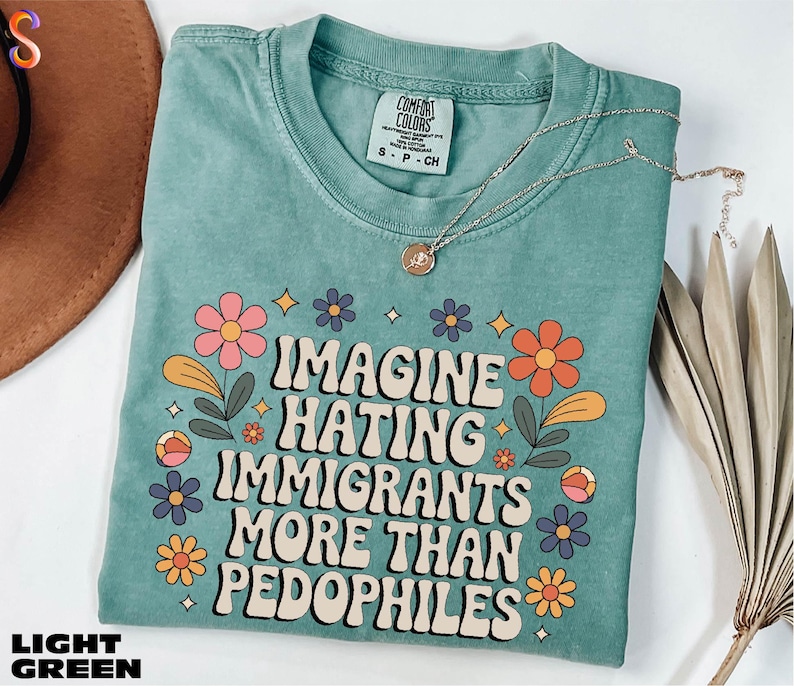 A turquoise t-shirt with a colorful graphic design and text, along with a necklace and some dried leaves, on a white background.