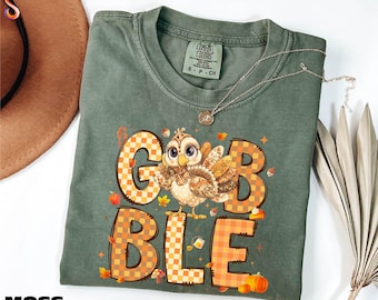 May include: Moss green t-shirt with a turkey graphic and the word "GOBBLE" in orange and brown checkered letters. The turkey has large eyes and is surrounded by fall-themed illustrations. A silver necklace is draped across the shirt.