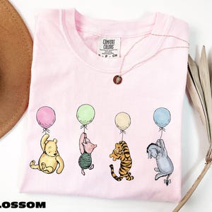 A pink t-shirt with illustrations of four cartoon characters from the Winnie the Pooh series, including Tigger, Tigger's friend, Tigger, and Eeyore, each holding a balloon. The t-shirt is displayed on a white background, with a brown hat and a plant visible in the background.