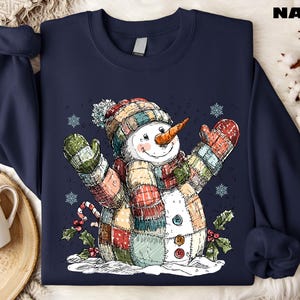 May include: Navy blue sweatshirt featuring a whimsical snowman illustration. The snowman is adorned with a patchwork scarf, hat, and mittens in various colors. A carrot nose and button details complete the design. The word "NAVY" is printed at the top.