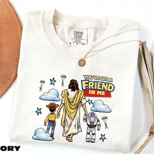 a white t-shirt with a cartoon illustration of a person holding hands with two children, one of whom is a character from the Toy Story franchise. The text on the t-shirt reads "You've got a Friend in Me".