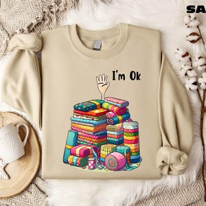 May include: A beige sweatshirt with a cartoon hand reaching up and the text "I'm Ok". The hand is surrounded by a stack of colorful fabric scraps.