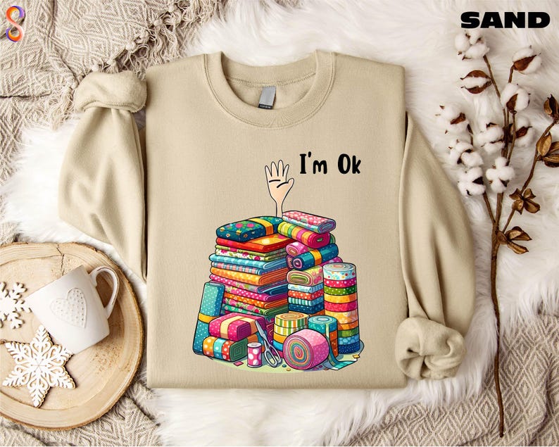 May include: A beige sweatshirt with a cartoon hand reaching up and the text "I'm Ok". The hand is surrounded by a stack of colorful fabric scraps.
