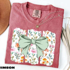 a red t-shirt with a bow design, placed on a white surface alongside a hat and some dried leaves.