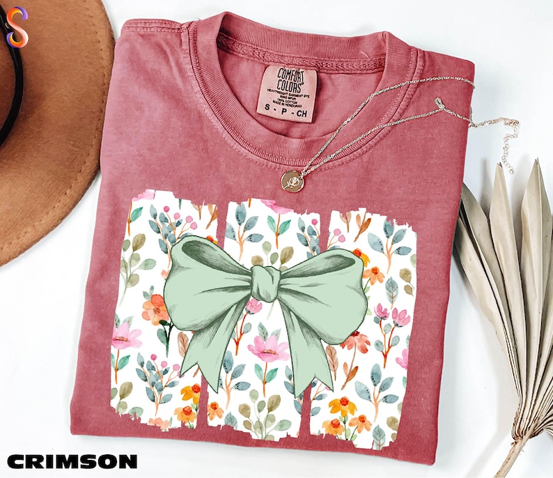 a red t-shirt with a bow design, placed on a white surface alongside a hat and some dried leaves.