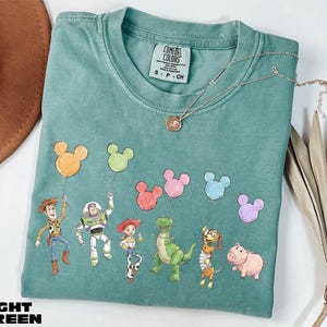A light green t-shirt with a design of various cartoon characters, including a cowboy, a girl, a dinosaur, a pig, and a piggy, along with a Mickey Mouse balloon. The t-shirt is placed on a white surface, and there is a brown hat and some dried leaves nearby.