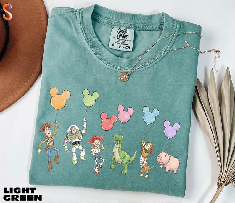 A light green t-shirt with a design of various cartoon characters, including a cowboy, a girl, a dinosaur, a pig, and a piggy, along with a Mickey Mouse balloon. The t-shirt is placed on a white surface, and there is a brown hat and some dried leaves nearby.