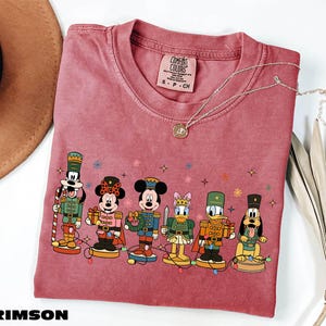 A pink t-shirt with a graphic of Disney characters, including Mickey Mouse, Minnie Mouse, Donald Duck, Goofy, and Pluto, along with a necklace and a hat.