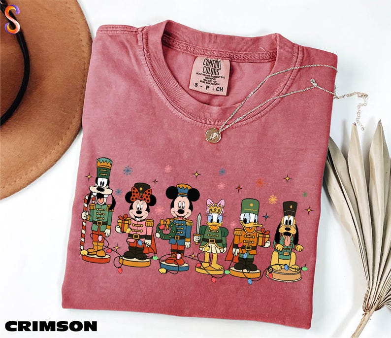 A pink t-shirt with a graphic of Disney characters, including Mickey Mouse, Minnie Mouse, Donald Duck, Goofy, and Pluto, along with a necklace and a hat.