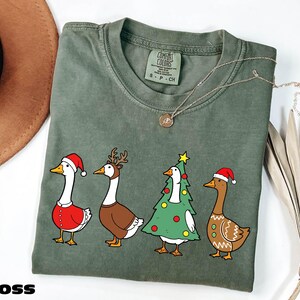 A green t-shirt with a Christmas-themed graphic of four cartoon ducks wearing Santa hats, along with a brown hat and some dried leaves.