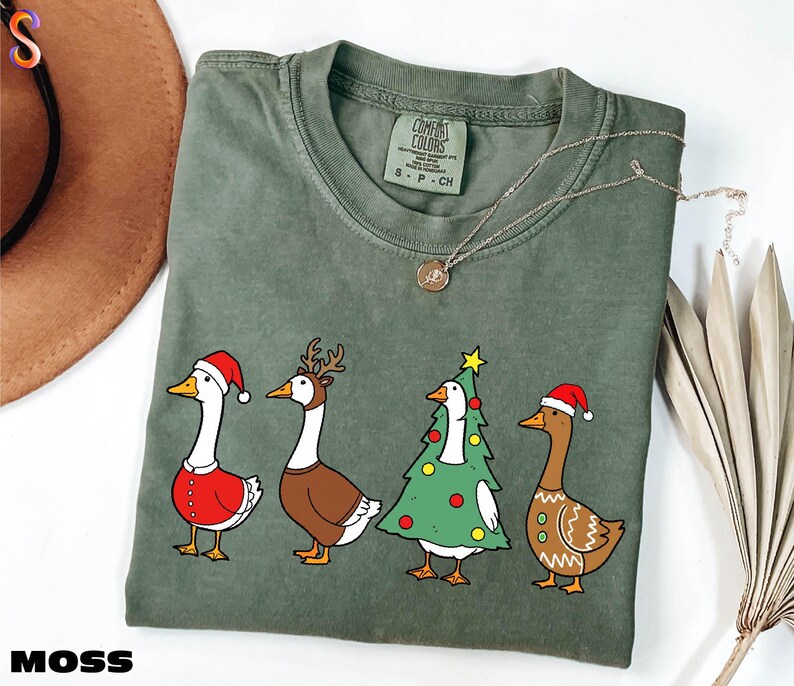 A green t-shirt with a Christmas-themed graphic of four cartoon ducks wearing Santa hats, along with a brown hat and some dried leaves.