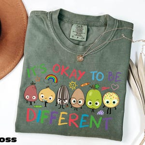 A green t-shirt with a colorful graphic design of cartoon characters and the text "It's OKAY TO BE DIFFERENT" printed on it.