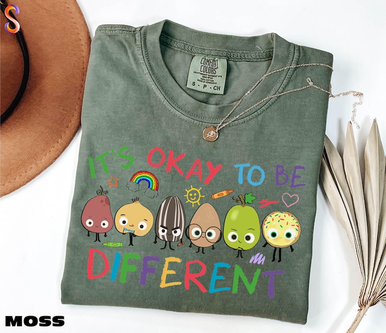 A green t-shirt with a colorful graphic design of cartoon characters and the text "It's OKAY TO BE DIFFERENT" printed on it.