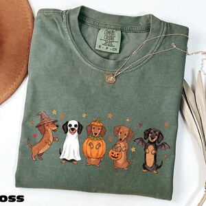 a green t-shirt with a Halloween-themed design of five dogs dressed in costumes, including a witch, a pumpkin, a bat, a dog with a pumpkin, and a dog with a bat.