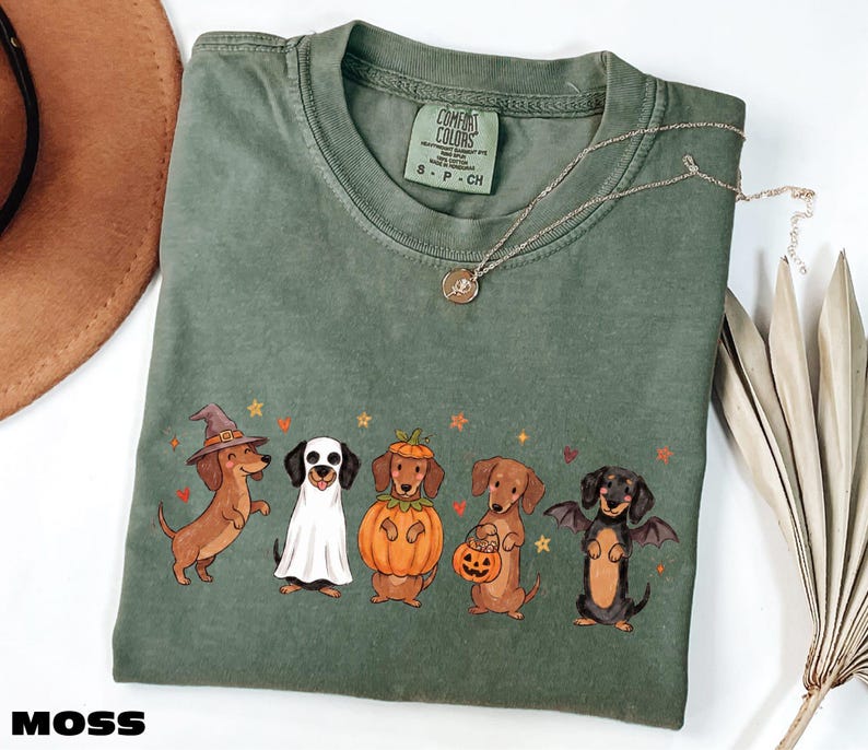 a green t-shirt with a Halloween-themed design of five dogs dressed in costumes, including a witch, a pumpkin, a bat, a dog with a pumpkin, and a dog with a bat.