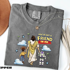 A gray t-shirt with a graphic design depicting a person in a yellow robe holding hands with two children, one of whom is holding a toy robot. The text on the t-shirt reads "You've got a friend in me".
