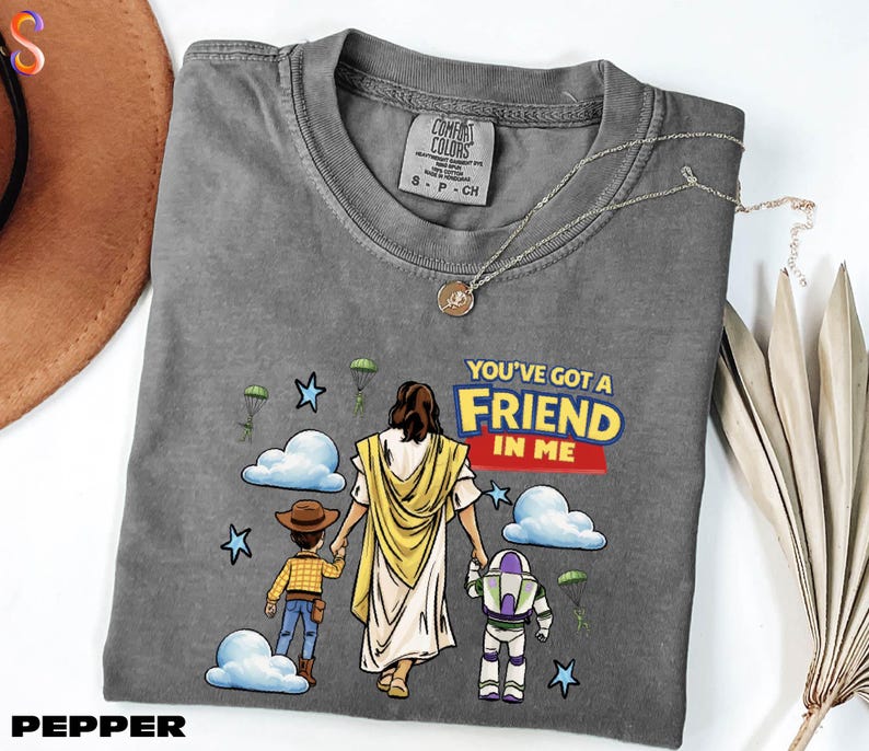A gray t-shirt with a graphic design depicting a person in a yellow robe holding hands with two children, one of whom is holding a toy robot. The text on the t-shirt reads "You've got a friend in me".
