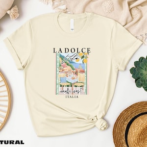 a beige t-shirt with a graphic design of a coastal town, along with the text "LADOLCE VITAMIN ITALIA". The t-shirt is displayed on a white background, surrounded by various items such as a straw hat, a woven basket, and a potted plant.