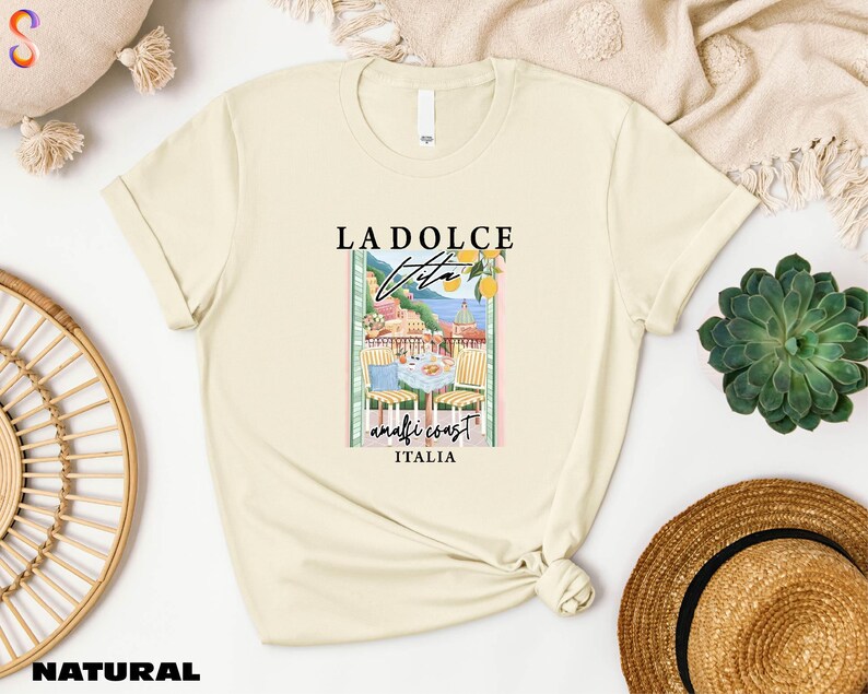 a beige t-shirt with a graphic design of a coastal town, along with the text "LADOLCE VITAMIN ITALIA". The t-shirt is displayed on a white background, surrounded by various items such as a straw hat, a woven basket, and a potted plant.
