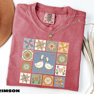 a red t-shirt with a colorful quilt-like design, including various geometric shapes, flowers, and birds. The t-shirt is placed on a white surface, with a brown hat and some dried leaves nearby.
