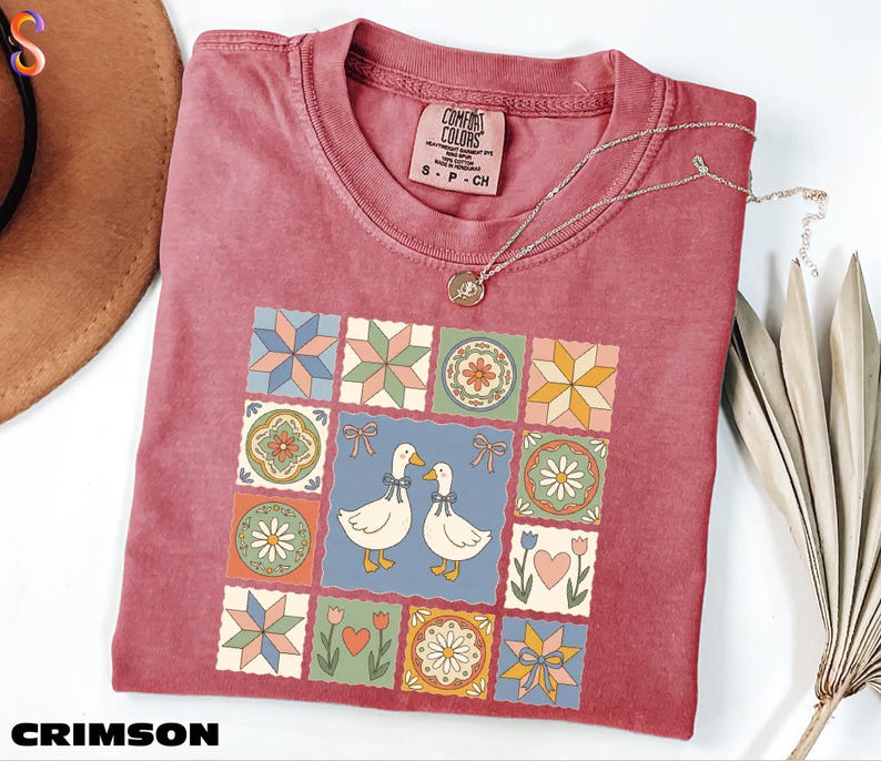 a red t-shirt with a colorful quilt-like design, including various geometric shapes, flowers, and birds. The t-shirt is placed on a white surface, with a brown hat and some dried leaves nearby.