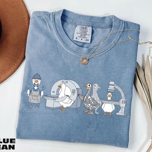 Könnte beinhalten: Blaues Jeans-T-Shirt mit einer Grafik von Cartoon-Gänsen in medizinischen Umgebungen. Eine Gans trägt einen Hut und eine Latzhose und hält Schilder mit "L" und "R". Andere Gänse befinden sich in der Nähe eines MRT-Geräts und eines Röntgengeräts.