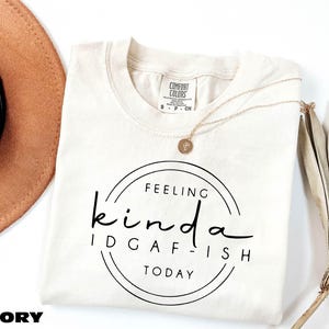 a white t-shirt with a black text design that reads "Feeling kinda idgafish today", accompanied by a brown hat and a plant.
