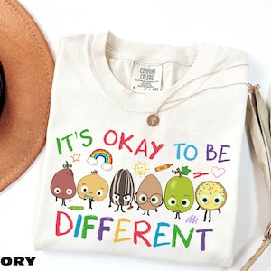 A white t-shirt with a colorful graphic design featuring a group of anthropomorphic fruits and vegetables, along with the text "It's OKAY TO BE DIFFERENT".