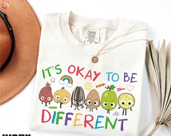 May include: Ivory-colored t-shirt with the text "IT'S OKAY TO BE DIFFERENT" in colorful letters. The shirt features cartoon illustrations of various food items with faces, including a pear, onion, and sunflower seed.