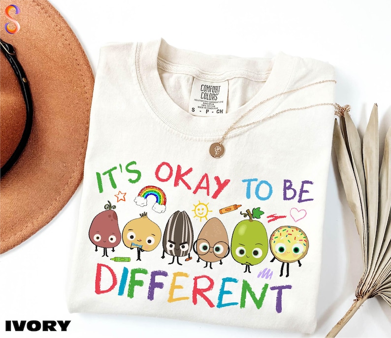 A white t-shirt with a colorful graphic design featuring a group of anthropomorphic fruits and vegetables, along with the text "It's OKAY TO BE DIFFERENT".