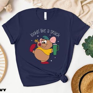 K&ouml;nnte beinhalten: Marineblaues T-Shirt mit einer Cartoon-Maus, die einen gr&uuml;nen Becher und K&auml;se h&auml;lt. Der Text "lookin' like a snack" befindet sich &uuml;ber der Figur. Ein l&auml;ssiges Kleidungsst&uuml;ck.