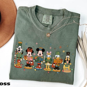 A green t-shirt with a graphic of Disney characters, including Minnie Mouse, Mickey Mouse, Donald Duck, Goofy, and Pluto, standing on a snowy surface.