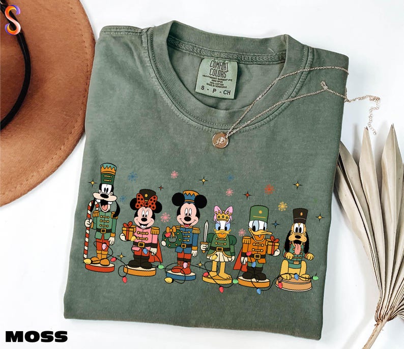 A green t-shirt with a graphic of Disney characters, including Minnie Mouse, Mickey Mouse, Donald Duck, Goofy, and Pluto, standing on a snowy surface.