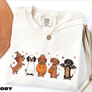 A white t-shirt with a graphic design of five dogs dressed in costumes, including a witch, pumpkin, bat, and dog with a pumpkin.