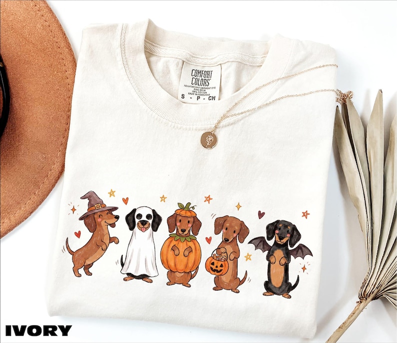 A white t-shirt with a graphic design of five dogs dressed in costumes, including a witch, pumpkin, bat, and dog with a pumpkin.