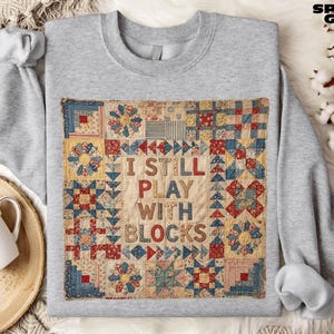 Peut inclure: Un sweat-shirt gris clair avec un motif patchwork coloré et le texte "I STILL PLAY WITH BLOCKS". Le patchwork comprend des motifs floraux et géométriques rouges, bleus et jaunes. Le sweat-shirt est étiqueté "SPORT GREY".
