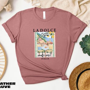 a pink t-shirt with a graphic design of a coastal town, along with the text "LADOLCE" and "Vite" printed on it. The t-shirt is displayed on a white background, surrounded by various items such as a straw hat, a woven basket, and a potted plant.