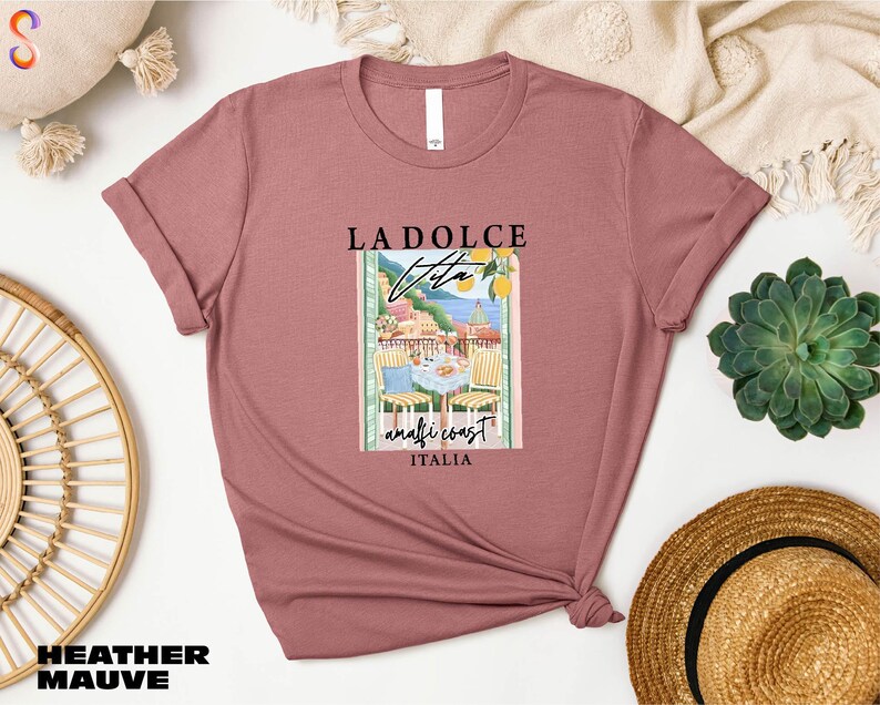 a pink t-shirt with a graphic design of a coastal town, along with the text "LADOLCE" and "Vite" printed on it. The t-shirt is displayed on a white background, surrounded by various items such as a straw hat, a woven basket, and a potted plant.