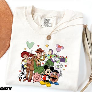 A white t-shirt with a colorful graphic of characters from the Toy Story movie, including Woody, Buzz Lightyear, Lassie, and other beloved characters.