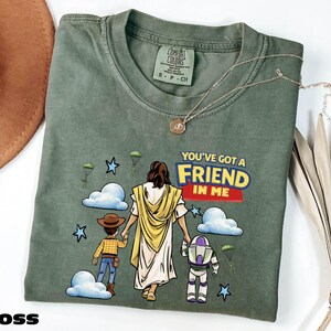 A green t-shirt with a cartoon illustration of a person holding a Bible and a child, along with the text "You've Got a FRIEND in Me" and a Bible verse reference.