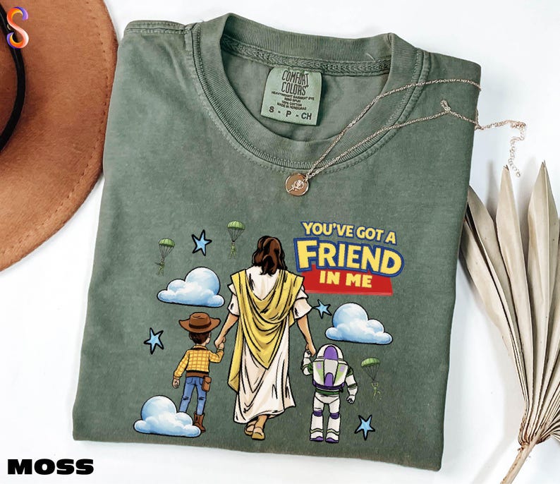 A green t-shirt with a cartoon illustration of a person holding a Bible and a child, along with the text "You've Got a FRIEND in Me" and a Bible verse reference.