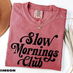 a pink t-shirt with the text "Slow Mornings Club" printed on it, along with a necklace and a hat.