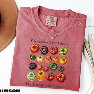 A pink t-shirt with a graphic of various colorful tomatoes, along with the text "Friends Don't Let Friends" and "Buy Store Bought Tomatoes" printed on it. The t-shirt is folded and placed on a white surface, with a brown hat and some leaves nearby.