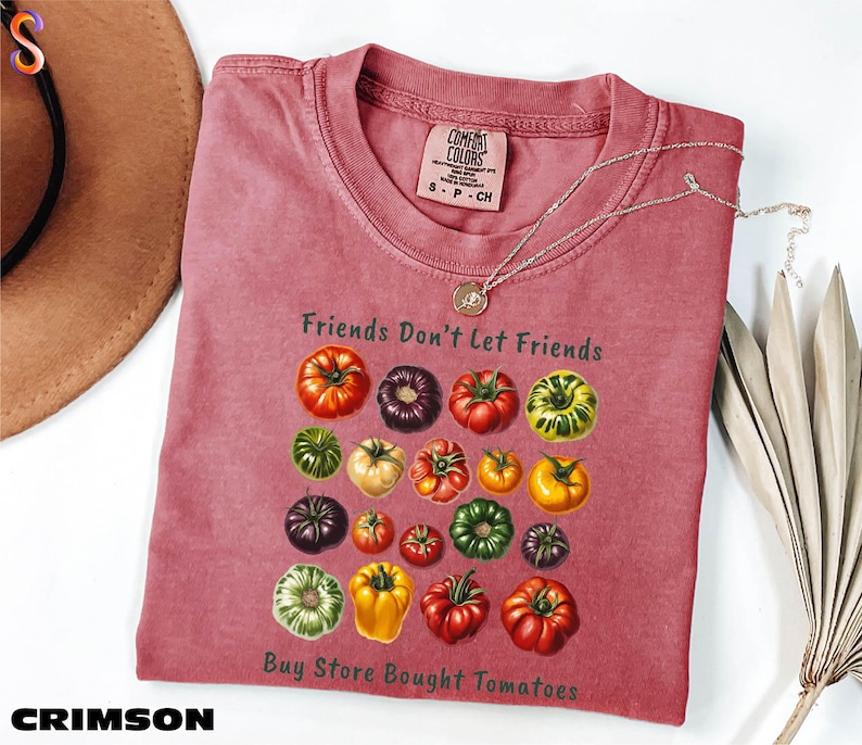 A pink t-shirt with a graphic of various colorful tomatoes, along with the text "Friends Don't Let Friends" and "Buy Store Bought Tomatoes" printed on it. The t-shirt is folded and placed on a white surface, with a brown hat and some leaves nearby.