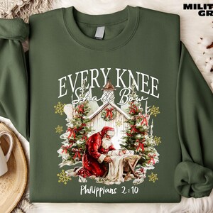 May include: Military green sweatshirt with a graphic of Santa kneeling before a manger, with Christmas trees and the text "Every Knee Shall Bow" and "Philippians 2:10". The sweatshirt is a crew neck style.