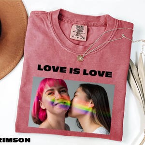 May include: Crimson-colored t-shirt with the text "LOVE IS LOVE" in black. The shirt features a photo of two people with rainbow-colored face paint. A Comfort Colors tag is visible on the collar.