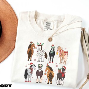 A white t-shirt with a graphic design of various horses wearing Christmas-themed outfits, including Santa hats and stockings. The t-shirt is placed on a white background, with a brown hat and a fan visible in the background.