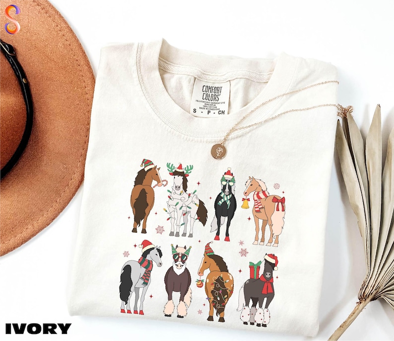 A white t-shirt with a graphic design of various horses wearing Christmas-themed outfits, including Santa hats and stockings. The t-shirt is placed on a white background, with a brown hat and a fan visible in the background.
